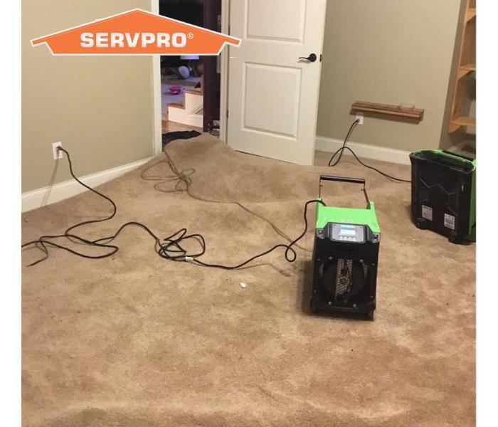 Two pieces of SERVPRO green quipment drying out brown carpet in a water damaged home. Orange SERVPRO house in corner